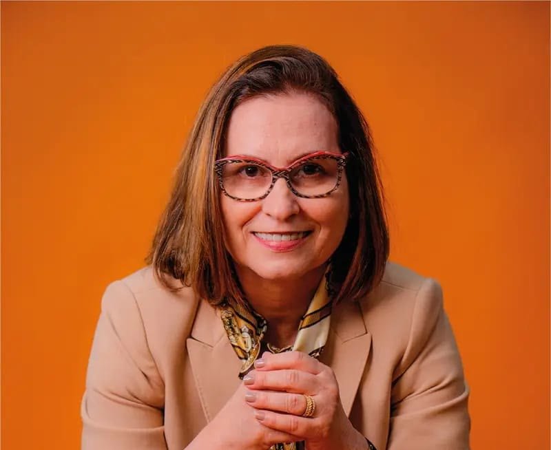 Alaide Bruno sorrindo, de blazer bege, em fundo laranja